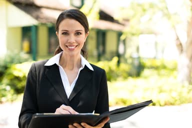 Woman wearing a suit and holding a portfolio with a house in the background Woman wearing a suit and holding a portfolio with a house in the background