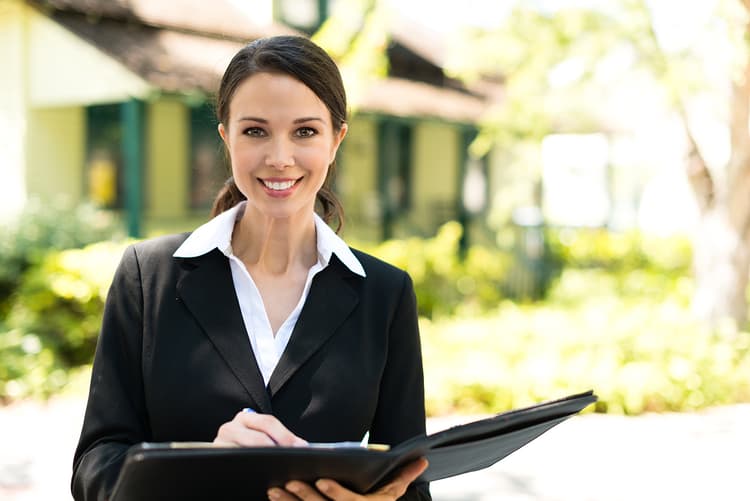 Woman wearing a suit and holding a portfolio with a house in the background Woman wearing a suit and holding a portfolio with a house in the background