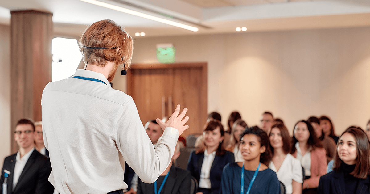 View from Behind of a Male Speaker Wearing a Headset Microphone Speaking to a Crowd Representing the Texas REALTOR® AE Seminar