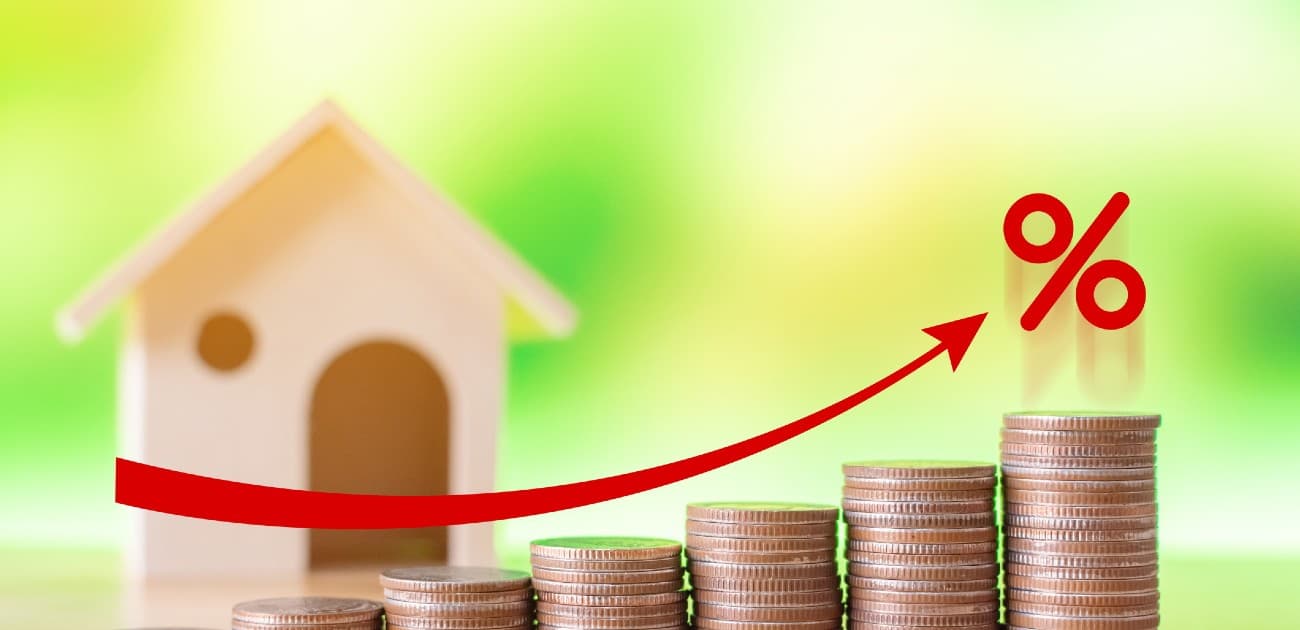 A picture of a house miniature behind a set of coin stacks, with a drawn-on red arrow curving upward toward a percent sign.