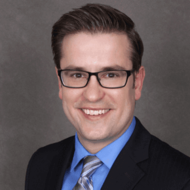 White man with brown hair and glasses smiling, wearing a suit with a blue shirt