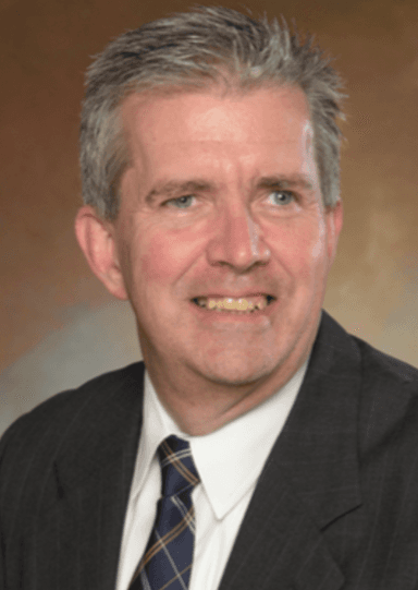 Photo of older white man in black suit with white shirt, he is smiling