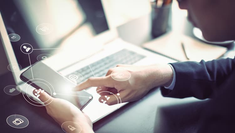 Man sitting at a desk and holding a cell phone with a laptop in the background Man sitting at a desk and holding a cell phone with a laptop in the background