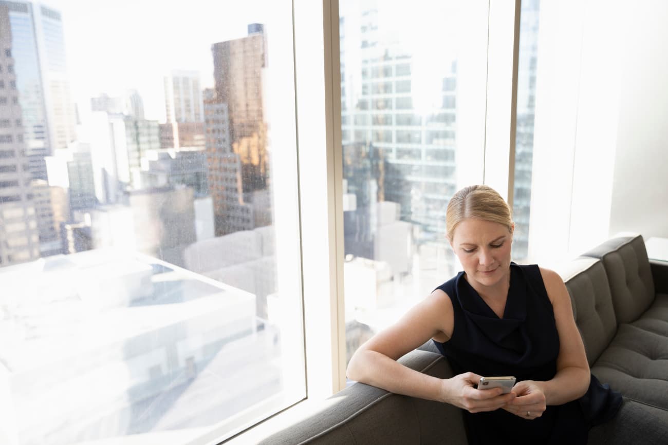 Woman sitting on a couch, next to a window, texting on her phone.