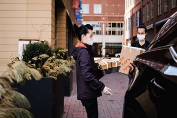 Woman and man wearing masks and gloves carrying a tray of food and a box practicing safe delivery