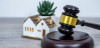 An auction gavel sits to the center right in the frame, with a small house miniature slightly behind it.